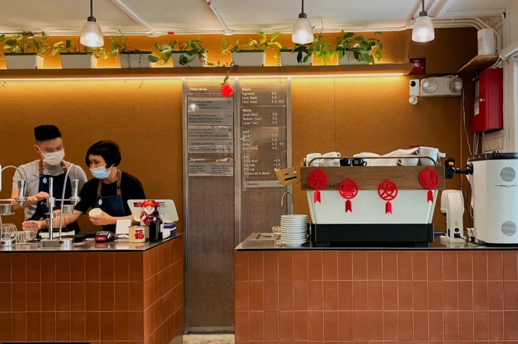 Baristas wearing masks behind a coffee counter, preparing drinks. The cafe interior features warm lighting, potted plants, and festive red decorations.