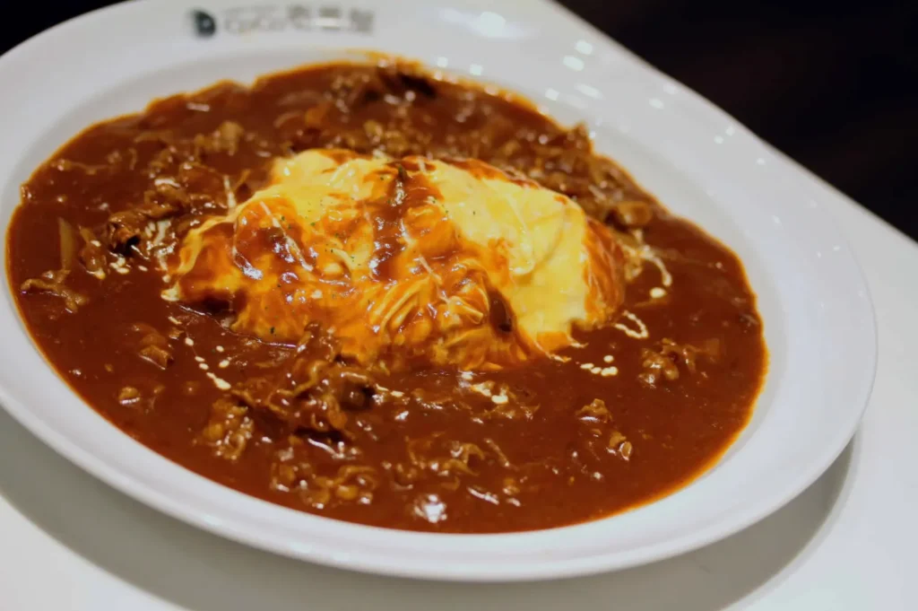 A plate of creamy omurice topped with rich, brown demi-glace sauce, creating a savory and indulgent presentation. The dish exudes a warm, comforting vibe.