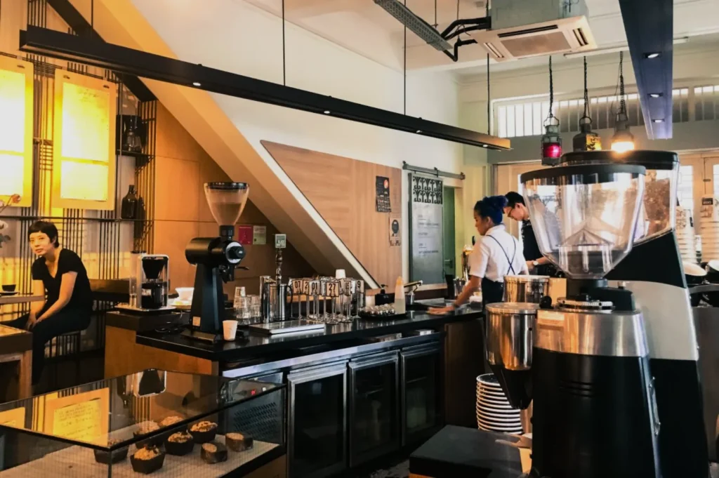 A cozy cafe with warm lighting. Two baristas work behind the counter with coffee equipment, while a woman sits at a table, creating a relaxed atmosphere.