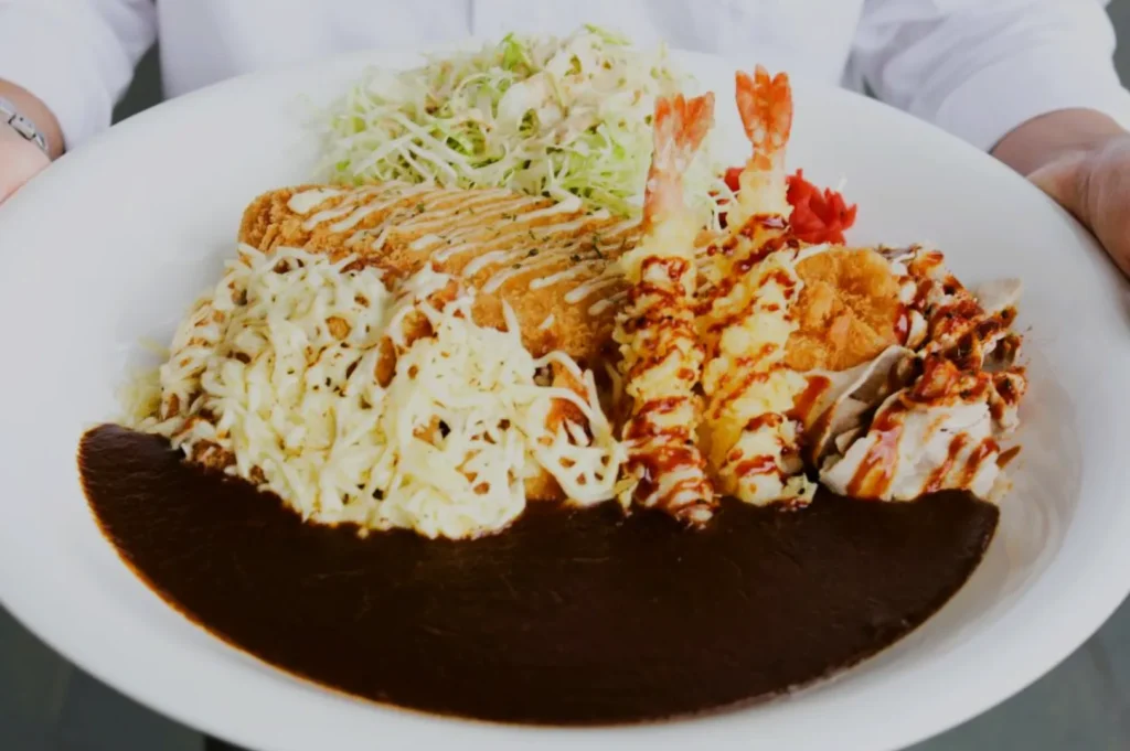 A large white plate holds a Japanese curry dish featuring crispy tempura shrimp, shredded cabbage, creamy sauce, and rich, dark curry. The presentation is vibrant and appetizing.