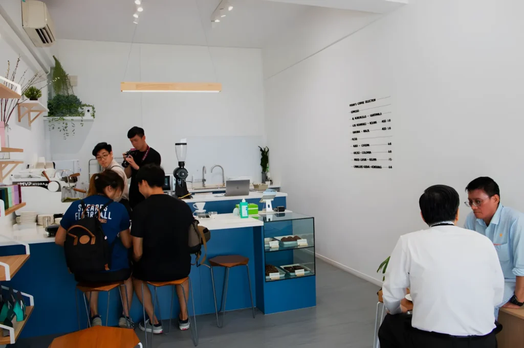 A modern coffee shop interior with a minimalist design. People sit on stools at a counter on the left, near a barista and an espresso machine. Two men chat at a small table on the right. The walls are white, with menu text and greenery for decor. The tone is casual and inviting.