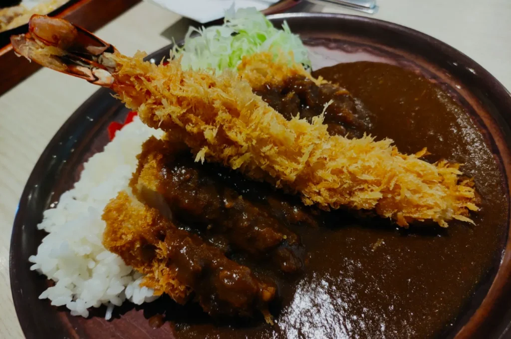 A plate of Japanese curry rice topped with crispy fried shrimp and shredded cabbage, creating a hearty and savory presentation with a warm, satisfying tone.