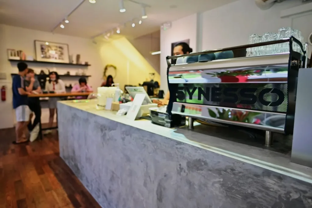 A modern cafe with a Synesso espresso machine on a concrete counter. People chat near wooden tables. Warm lighting creates a cozy, inviting atmosphere.
