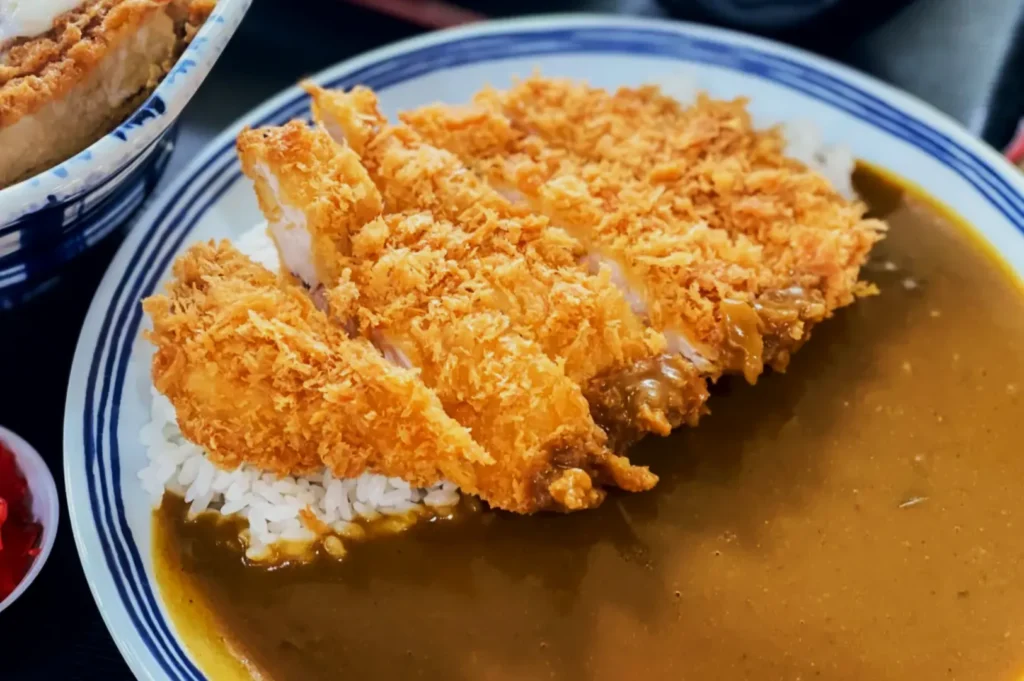 A crispy, golden-brown katsu lies on a bed of white rice, partially covered in rich, brown curry sauce. The dish sits on a striped blue plate.