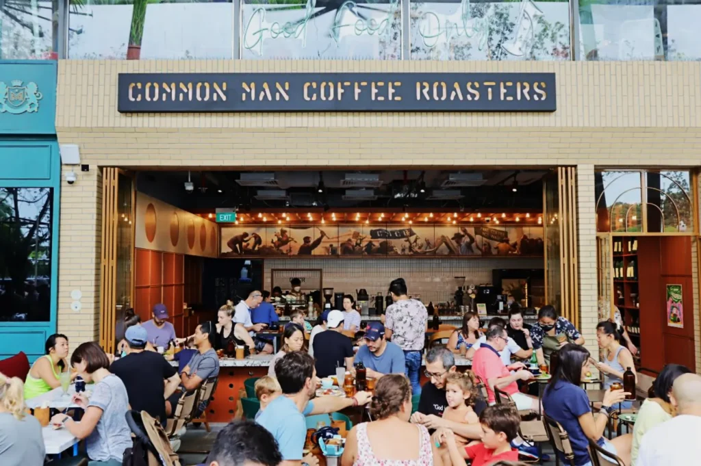 Bustling café scene at "Common Man Coffee Roasters." Patrons fill outdoor tables, enjoying drinks and food, creating a lively, social atmosphere.