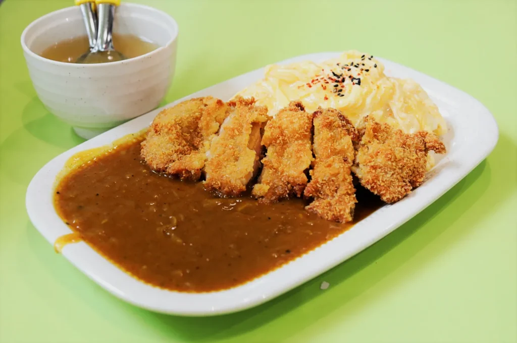 A plate of Japanese curry with crispy breaded cutlets, creamy egg topping, and rice, garnished with sesame seeds. A bowl of soup is beside it.