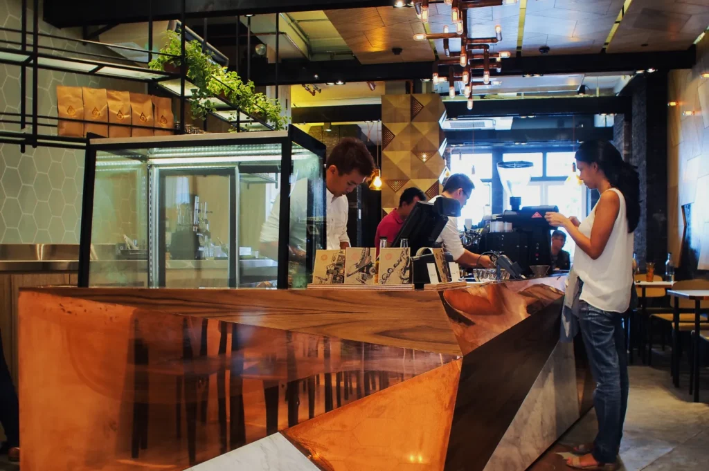Modern cafe interior with angular wooden counter, barista making coffee, and a woman ordering. Warm lighting, plants overhead, relaxed atmosphere.