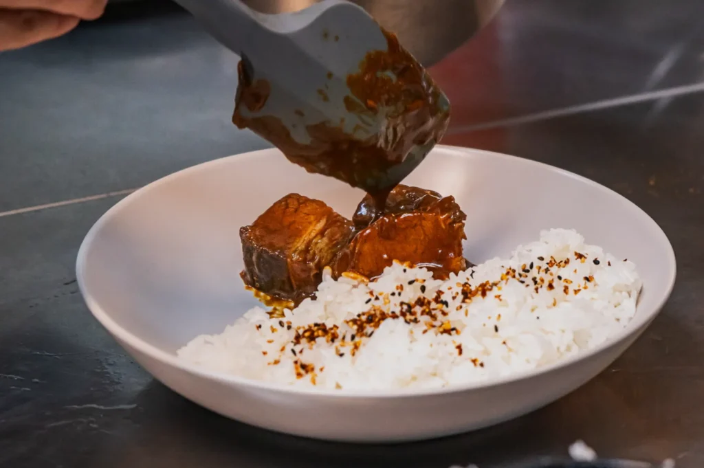 A person pours rich brown sauce over tender pork pieces next to seasoned white rice on a plate, creating a warm, appetizing meal presentation.