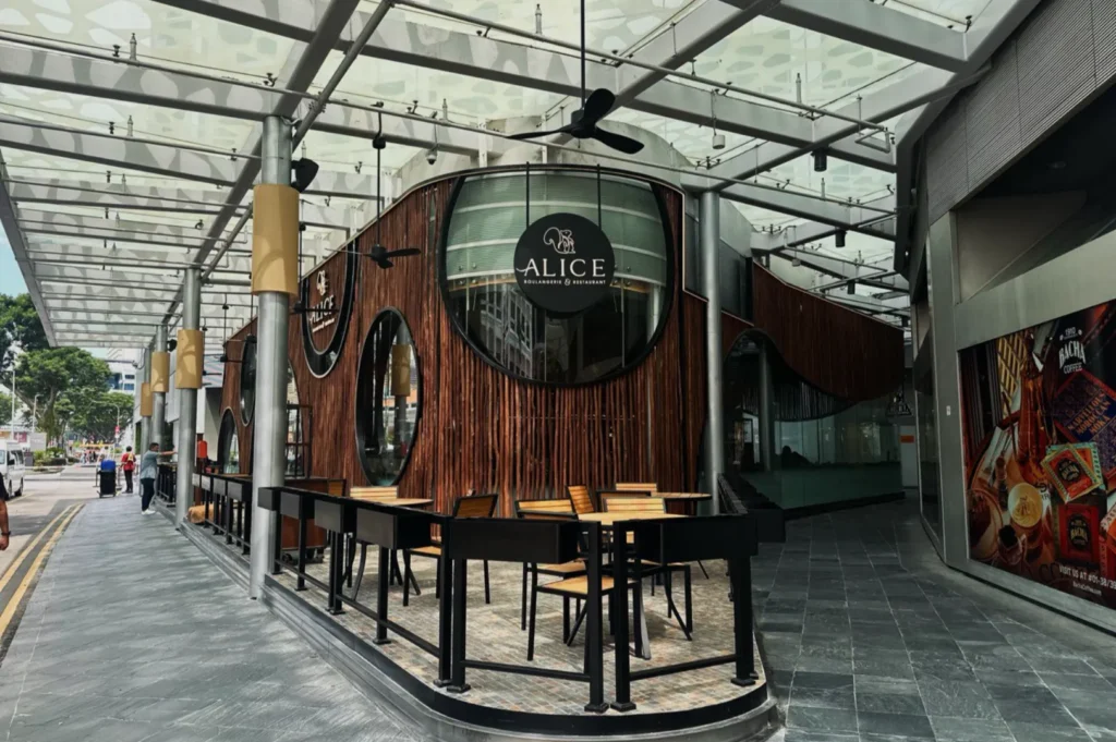 A stylish cafe with circular windows and wooden paneling, under a transparent canopy. Tables and chairs are set outside, creating a welcoming vibe.