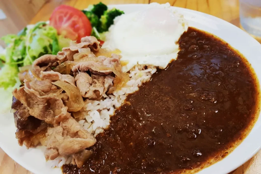 A plate with beef and onions over rice, topped with a fried egg. A rich brown curry sauce is poured on one side, with salad and tomato slices on the side.