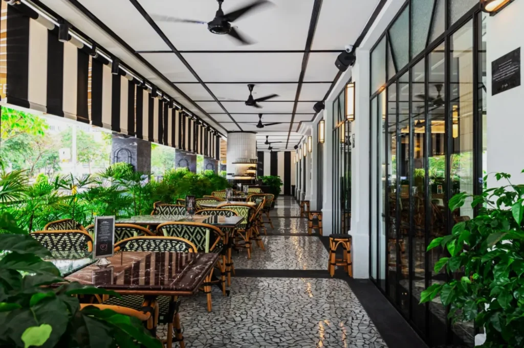 Spacious outdoor cafe with a black and white color scheme, featuring patterned tables, wicker chairs, ceiling fans, and lush green plants for a relaxed ambiance.