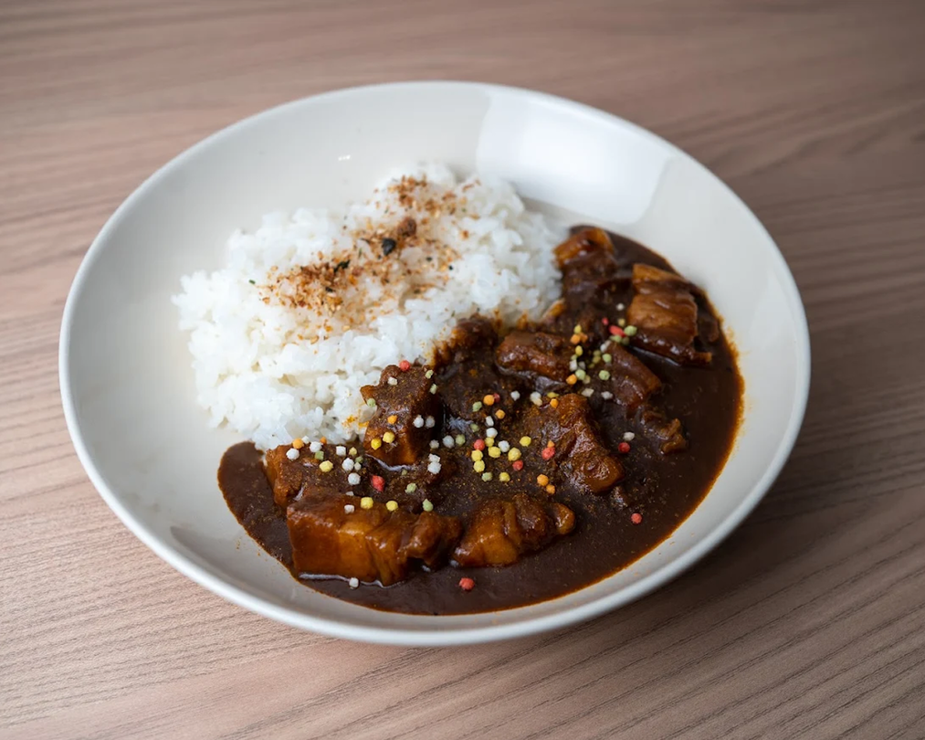 A close-up view of a rich, black curry sauce envelops tender short ribs, served alongside fluffy rice. This dish showcases the depth of flavors typical of Japanese curry rice, highlighting its hearty and satisfying nature.