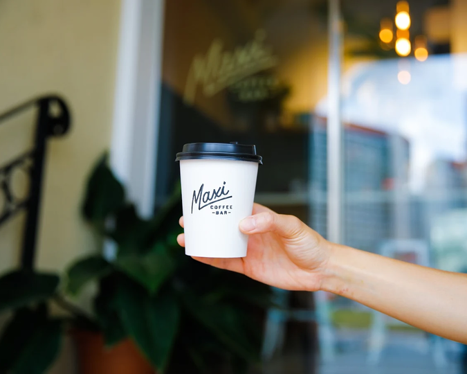 A close-up of a hand holding a cup of coffee, with the signature Maxi Coffee Bar backdrop softly blurred behind for emphasis