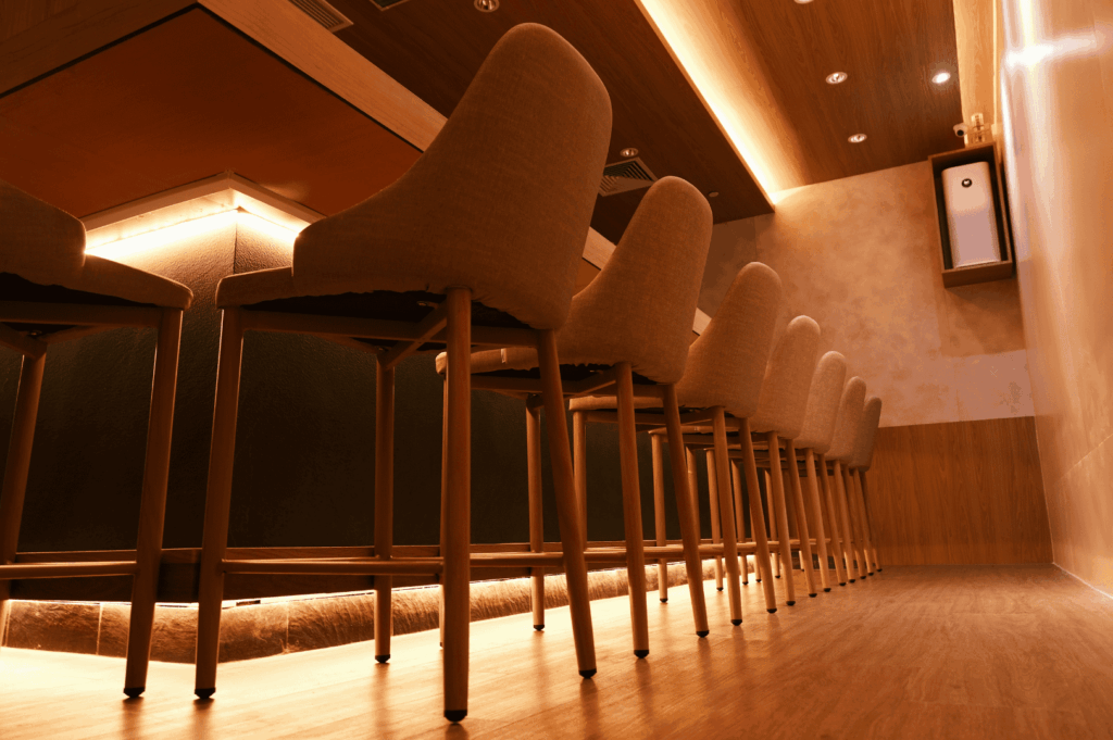 A row of high-backed bar stools lines a sleek wooden counter in a softly lit, modern dining space at Sushi Masa—warm tones and clean lines evoke quiet luxury and intimate omakase moments.