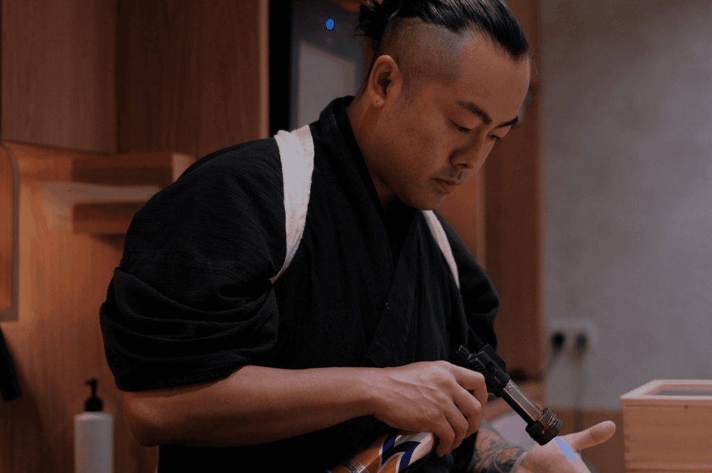 Chef Masa sears ingredients with a culinary torch at a sleek wooden counter, his focused technique embodying the quiet intensity and precision behind Sushi Masa by Ki-setsu’s omakase experience.