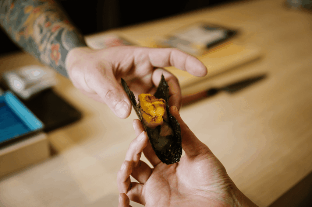 Chef Masa’s tattooed hand passes a uni-topped sushi to a diner across a wooden counter—an intimate gesture that captures the personal artistry and quiet reverence of the omakase experience at Sushi Masa by Ki-setsu.