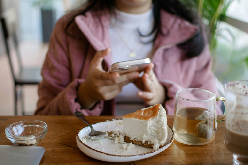 A person is intently reading restaurant reviews on their smartphone, with an array of colorful desserts, including cakes and pastries, elegantly displayed in the background. This scene captures the essence of a dessert lover's paradise, perfect for those seeking sweet treats in vibrant cafes along Orchard Road.