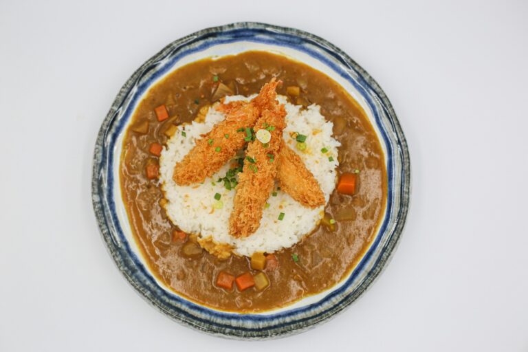 A steaming bowl of Japanese curry rice features a golden katsu cutlet resting atop fluffy white rice, accompanied by rich curry sauce, creating a satisfying meal that highlights the delicious flavors of Japanese cuisine. The dish is garnished with vibrant vegetables, adding to its hearty appeal.