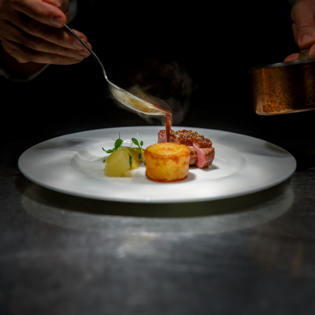 A chef carefully pouring warm soup onto a plated dish, highlighting precision, balance, and the artistry of fine cooking.