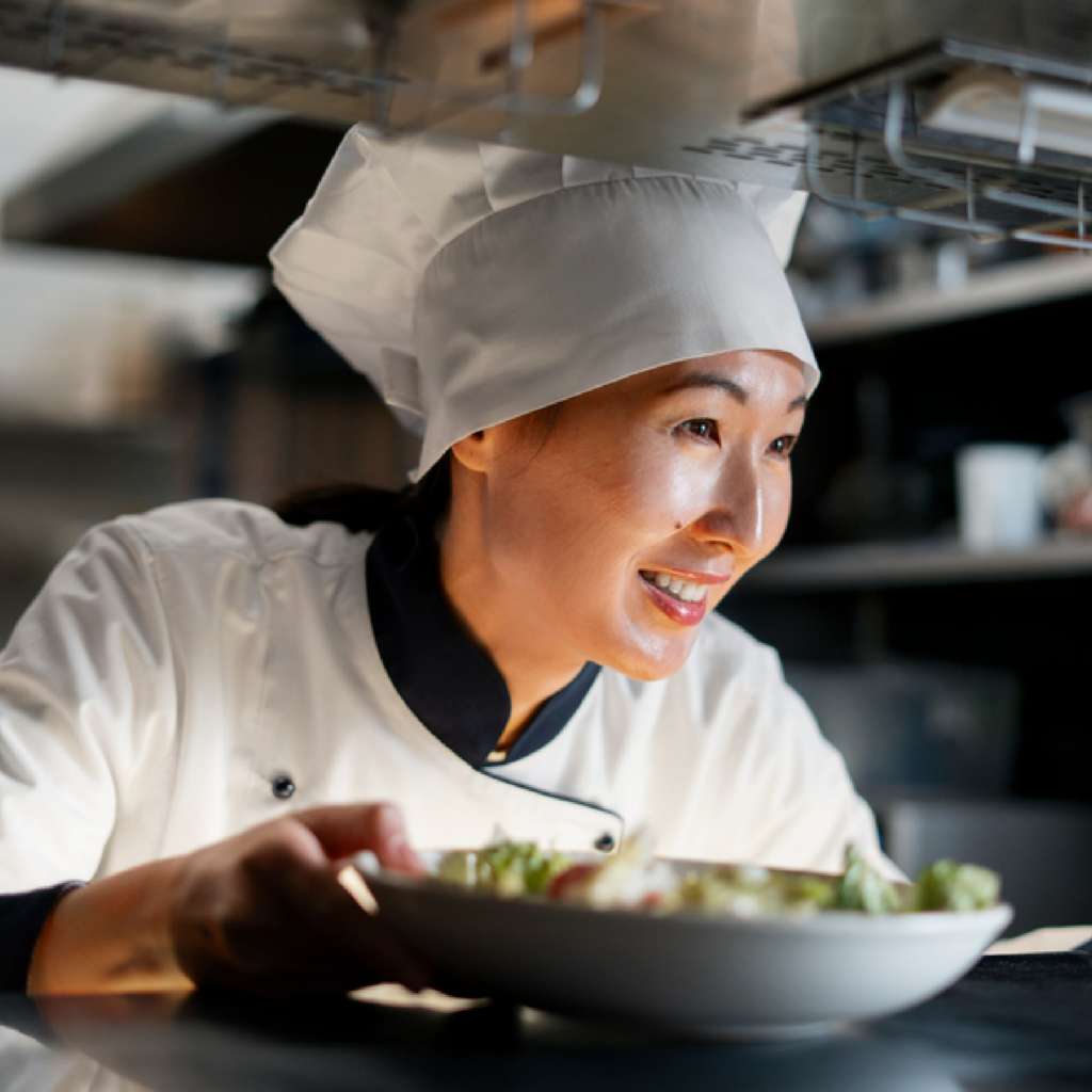 A woman chef gently placing a finished dish on the table, reflecting pride, craftsmanship, and attention to detail after cooking.