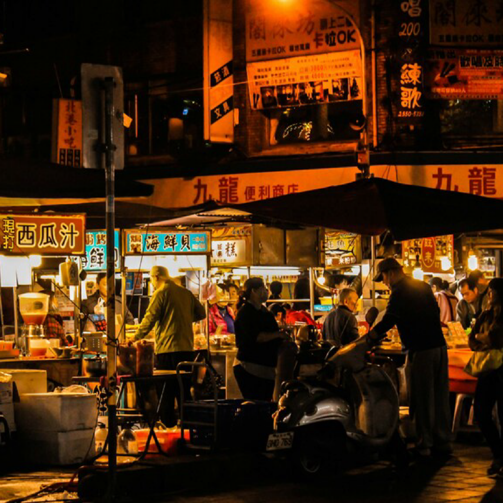 People browsing and purchasing food at a lively night market, surrounded by vibrant stalls, warm lights, and a bustling atmosphere.