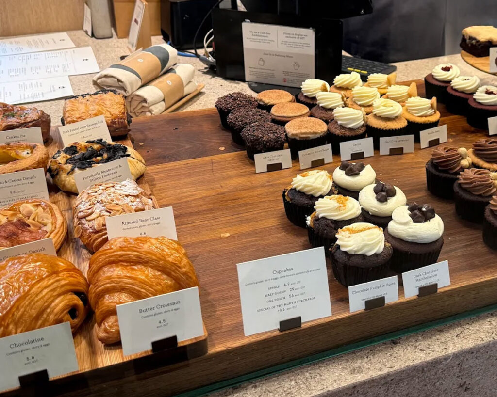 A dark chocolate cupcake topped with glossy ganache frosting, placed on a simple plate with a softly blurred bakery counter in the background.