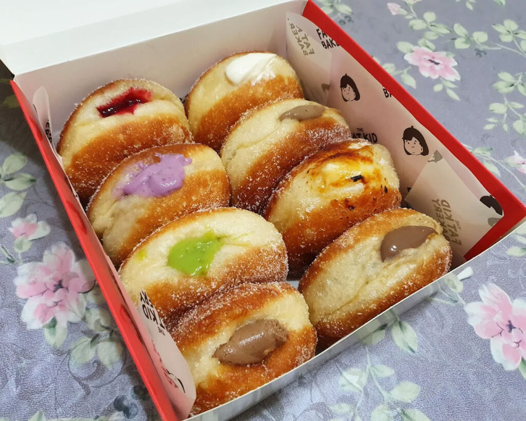 A golden sourdough bomboloni filled with cream and dusted with sugar, displayed on a plate with a café background featuring warm colours and casual seating.