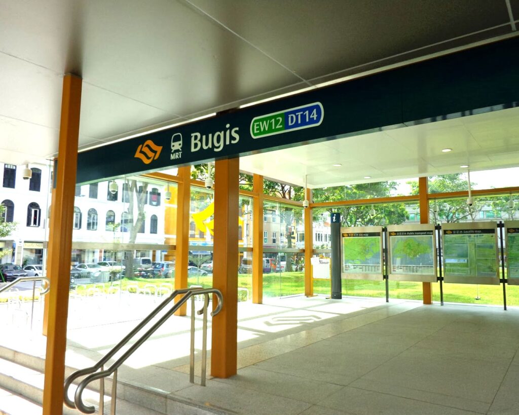 The entrance of Bugis MRT Station with its signboard visible, showing commuters walking in and out and the surrounding urban environment.