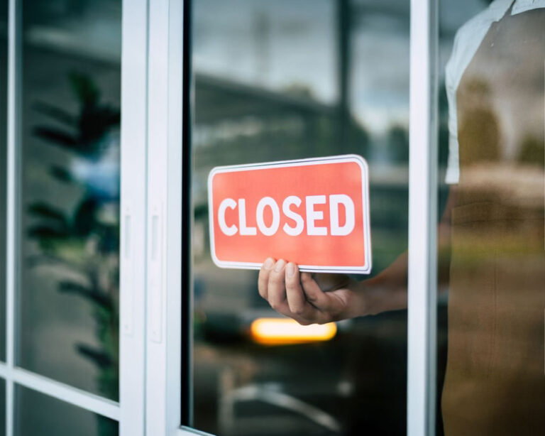 A close-up of a ‘Closed’ sign hanging on a glass restaurant door, reflecting a quiet interior and signalling the restaurant is temporarily not open to the public