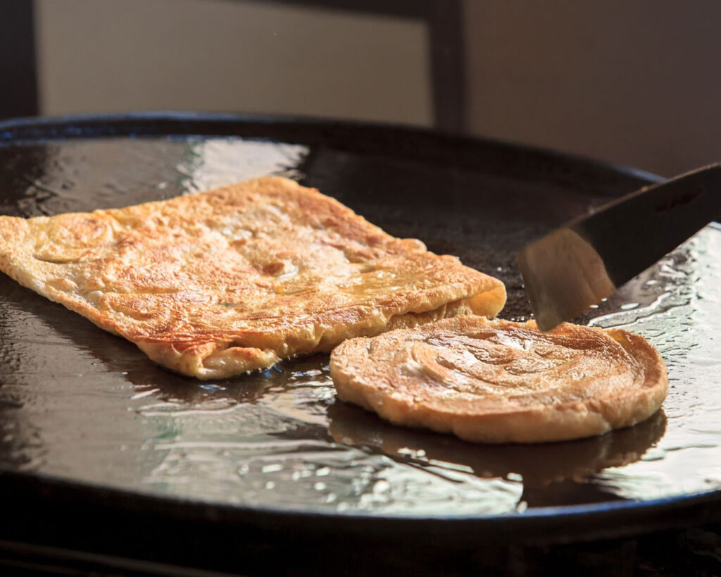 Parata cooking on a hot griddle, slowly turning golden and crisp as heat transforms the dough into flaky perfection.