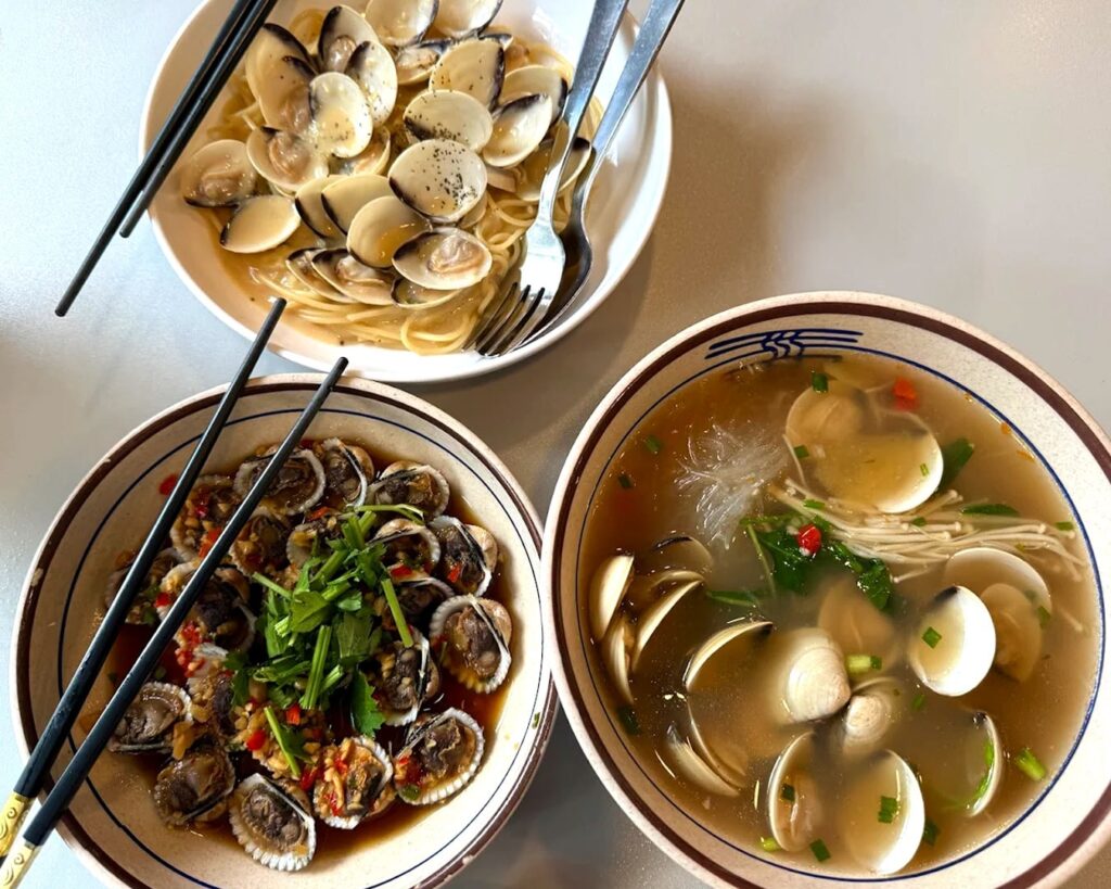 A bowl of lala bee hoon served on a wooden table at Fortune Centre, featuring clear broth, fresh clams, vermicelli noodles, and garnished with herbs, ready to be enjoyed.