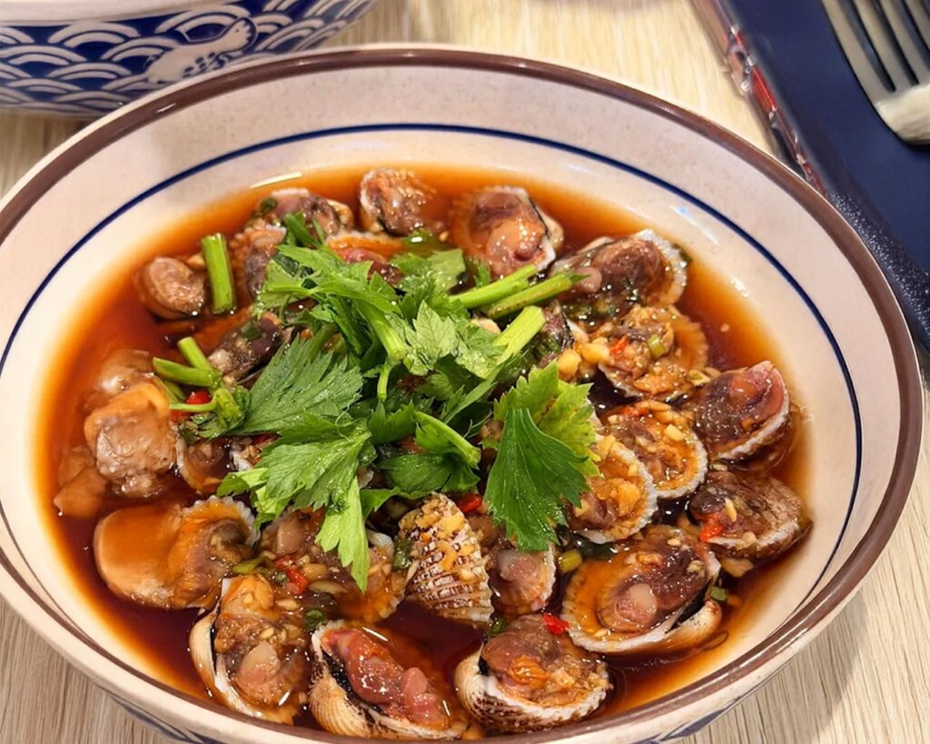 A tray of live clams and fresh cockles displayed in a wet market setting, showing their natural shells and freshness before cooking.
