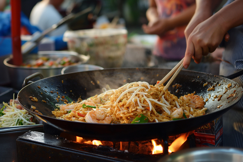 A cook’s hand stir-frying char kway teow in a hot wok, the flat rice noodles glistening with oil as steam rises, showing the intensity and skill behind the cooking process.