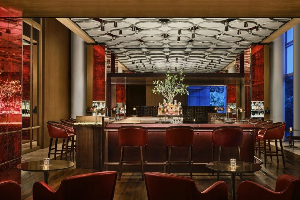 Luxurious bar interior with a modern design. Red leather chairs and warm lighting create an inviting atmosphere. Elegant ceiling fixtures and a central bar with bottles on display add sophistication.