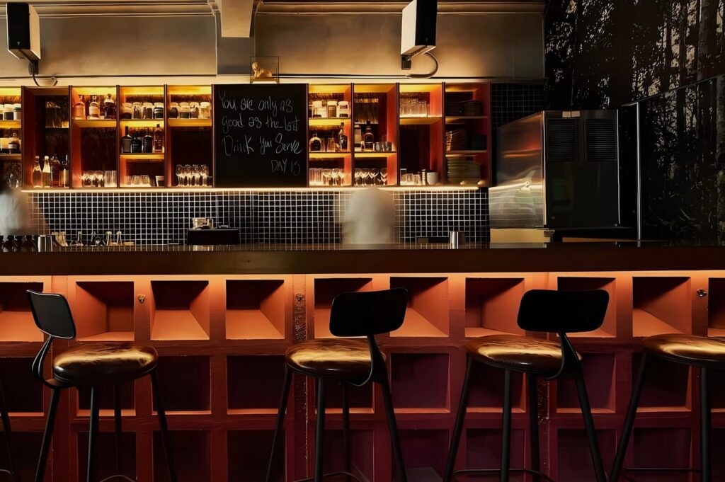 Stylish bar interior with high stools, a tiled bar counter, and warm lighting. Shelves with bottles and glassware. A chalkboard displays a message. Cozy ambiance.