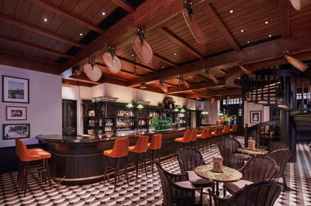 Stylish pub interior with a dark wood bar, orange bar stools, and a checkered floor. Ceiling fans and spiral staircase add vintage charm. Cozy and inviting atmosphere.