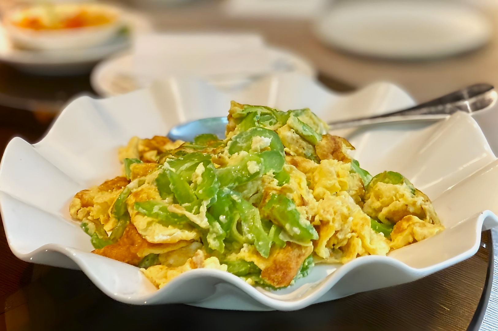 A white scalloped dish filled with a stir-fried egg and green bell pepper mix. The fork rests on the side. The setting is a casual dining table.