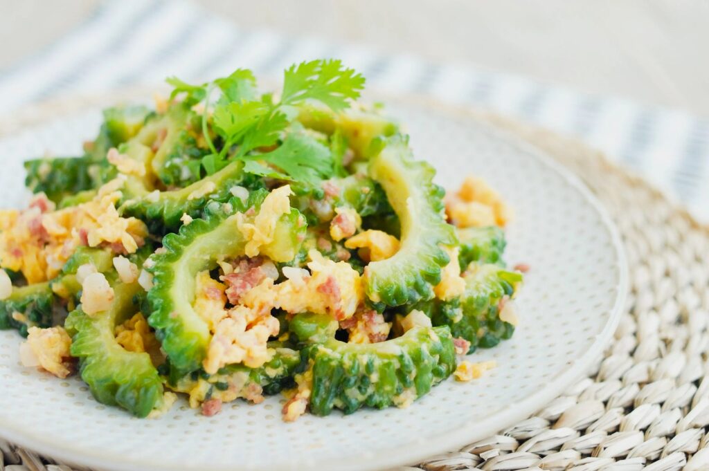 A beige plate with a stir-fried dish of bitter melon, scrambled eggs, and minced meat, garnished with fresh cilantro. A woven placemat and striped cloth in the background.