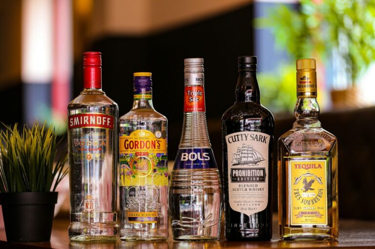 A row of five liquor bottles, including vodka, gin, triple sec, whisky, and tequila, stand on a wooden table beside a small potted plant, with a blurred indoor background.