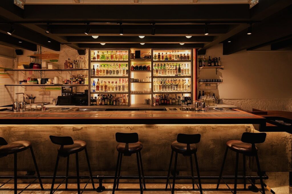 Dimly lit bar with a warm ambiance. Shelves stocked with diverse bottles glow behind a sleek counter. Five bar stools are evenly spaced in the foreground.