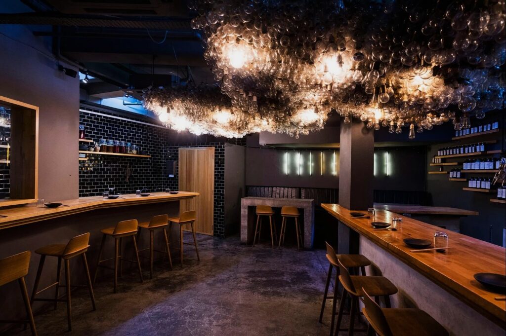 Stylish, dimly lit bar interior with wooden counters and stools. Unique ceiling covered with hanging lights creates a cozy, modern ambiance.