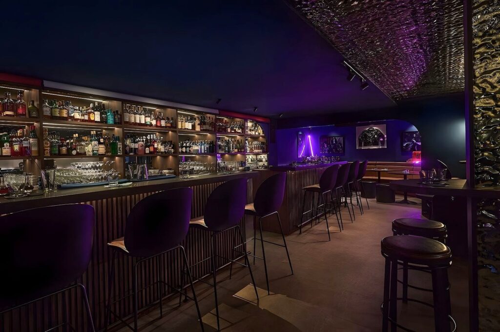 Dimly lit modern bar with purple accents, lined with high-backed stools. Shelves behind the counter display a variety of bottles, creating a cozy, intimate ambiance.