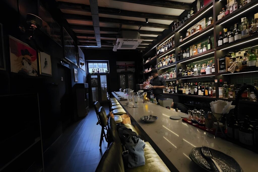 Dimly lit bar with a long counter, high shelves filled with various bottles, and a bartender preparing drinks. The atmosphere is cozy and intimate.