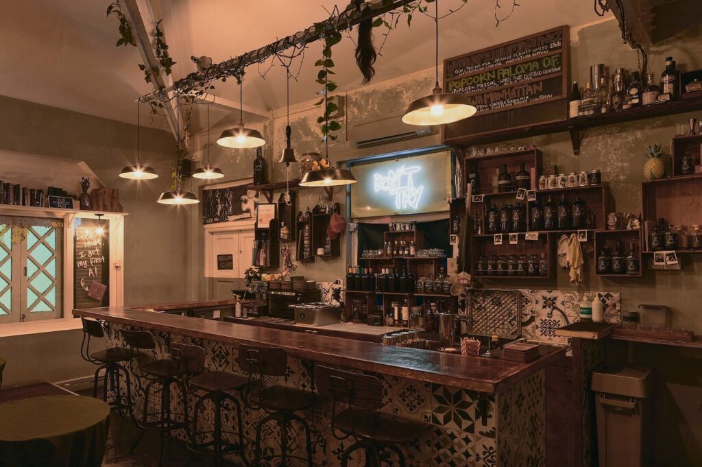 A cozy, dimly lit bar with vintage decor. Warm lights hang over a wooden counter with stools. Shelves hold various bottles, and a neon sign reads "Don't Try."