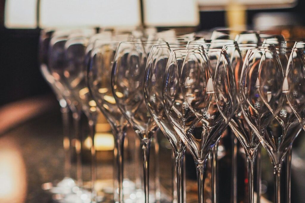 A close-up of multiple empty wine glasses arranged in rows, reflecting a warm light. The background is softly blurred, creating an elegant atmosphere.