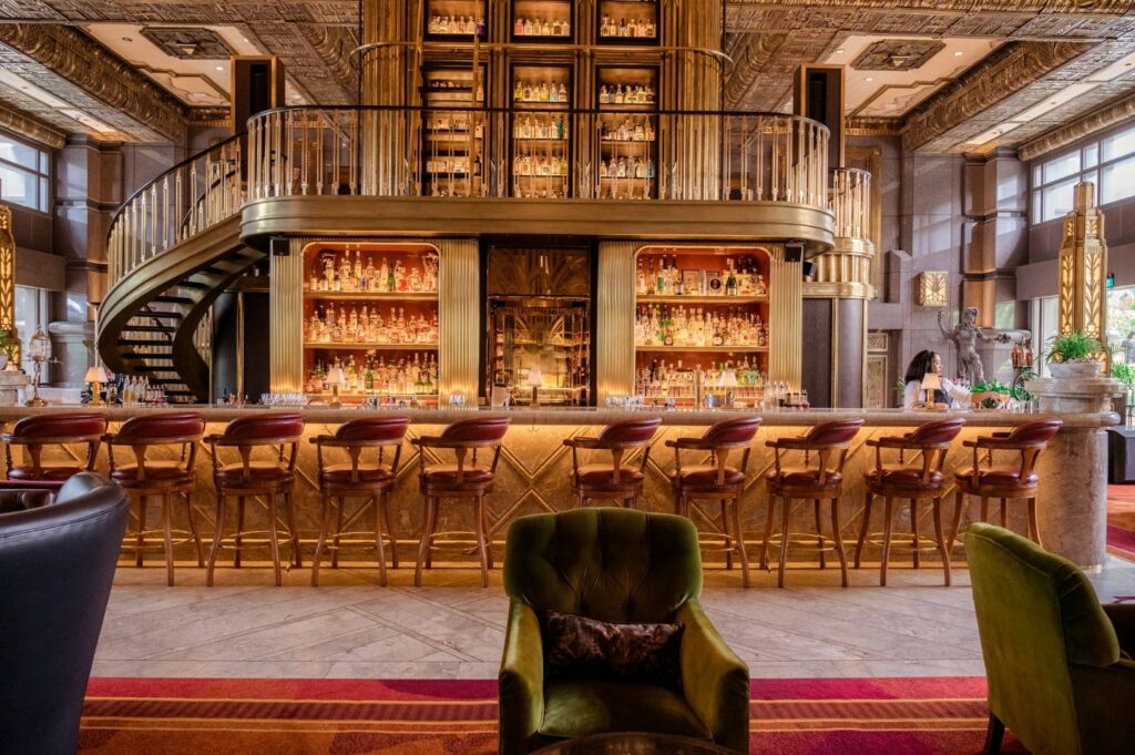 Luxurious bar with golden shelves filled with bottles, central spiral staircase, high-backed chairs, plush green armchairs, warm and elegant ambiance.