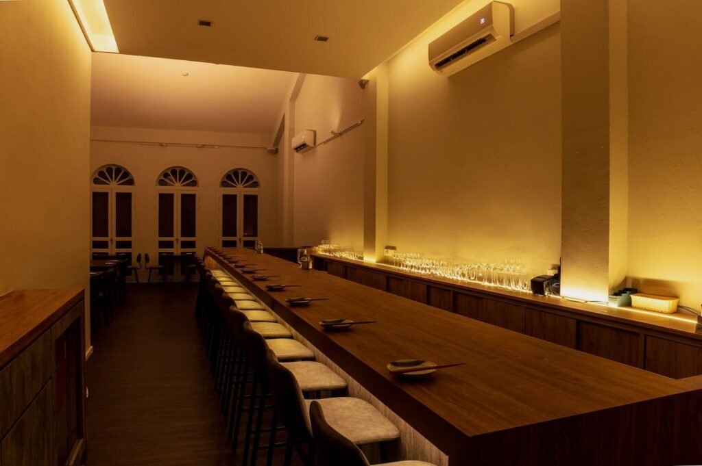 A cozy, dimly lit restaurant interior features a long wooden table with cushioned chairs, table settings, and ambient lighting. Arched windows add elegance.