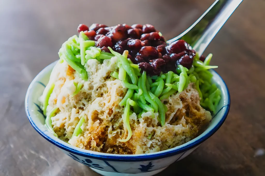 A bowl of shaved ice topped with vibrant green cendol and red beans, served with a shiny metal spoon. The image conveys a refreshing, sweet dessert.