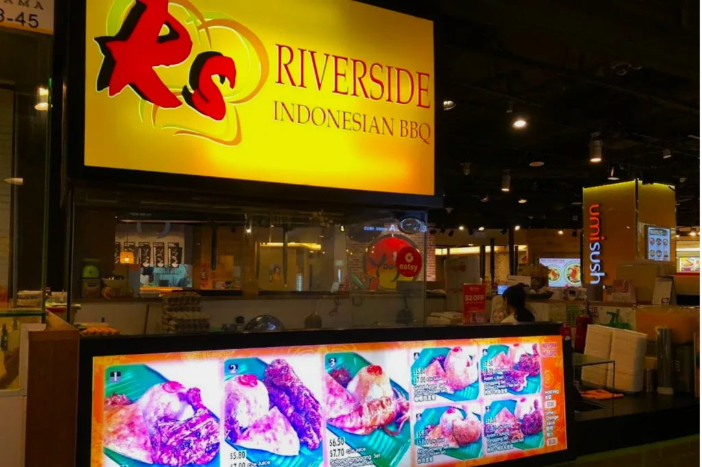 Illuminated sign reads "Riverside Indonesian BBQ" above a counter. Bright menu displays various grilled dishes. Warm, inviting atmosphere.