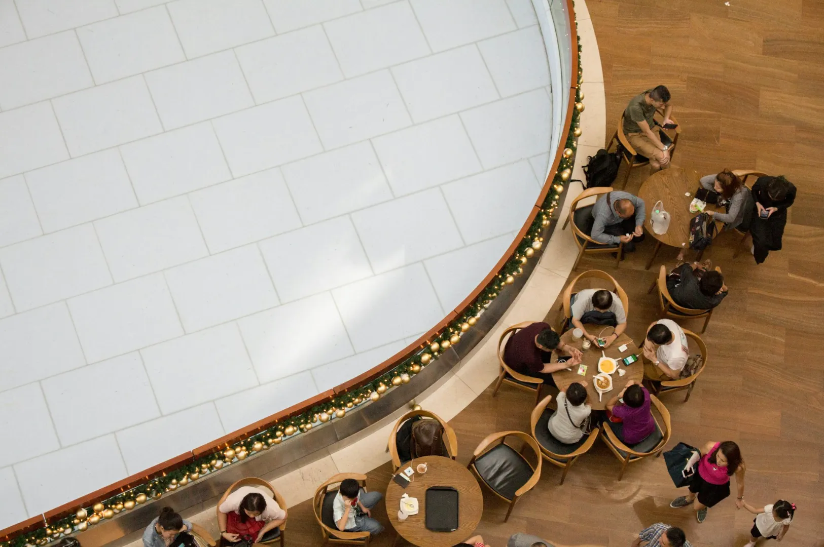 Overhead view of people sitting at a wooden-floored café with small round tables, adjacent to a large, decorative, white-tiled space, adorned with garland.